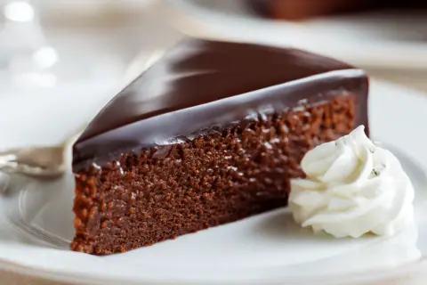 Ein Stück Sachertorte, wie im Sachertorte Rezept Original Lafer, mit glänzender Schokoladenglasur und einem Klecks Schlagobers auf einem weißen Teller.