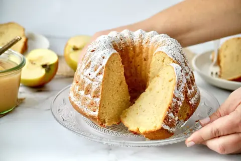 Saftiger Gugelhupf mit Puderzucker bestäubt, daneben Äpfel und ein Glas Apfelmus. Ein Stück Kuchen mit Apfelmus ist ein Genuss.
