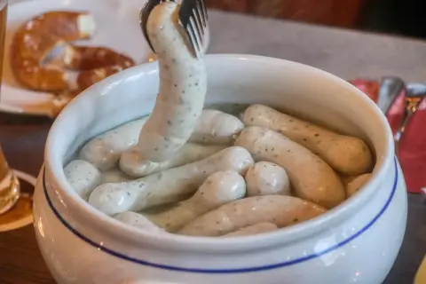 Eine Gabel hebt eine Weißwurst aus einem Topf, um sie zuzubereiten. Daneben liegt eine Brezel.