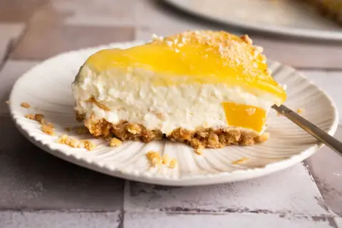 Ein cremiges Stück Kuchen ohne Backen mit gelber Fruchtschicht und knusprigem Boden auf einem Teller.