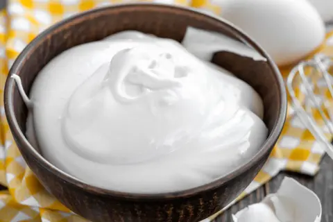 Perfekt geschlagener Eischnee in einer Schüssel, bereit für süße Kreationen. Daneben liegen Eier und ein Schneebesen.