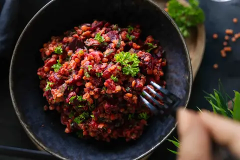 Ein herzhafter Linsen Rote Bete Salat, garniert mit Petersilie, wird mit einer Gabel aus einer dunklen Schüssel genommen.