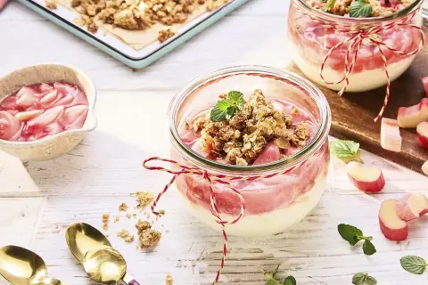 Zwei Gläser mit Rhabarber Crumble, garniert mit Minze. Frischer Rhabarber liegt daneben.