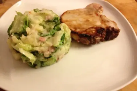 Ein gebratenes Schweinekotelett liegt neben einem Berg aus Kartoffelpüree mit Speck und **endivien untereinander**.