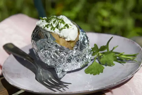 Folienkartoffeln vorkochen: Eine Kartoffel in Alufolie, belegt mit Quark und Kräutern, auf einem Teller mit Gabel und Petersilie.