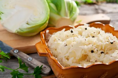 Schüssel mit Sauerkraut, Gewürzen und frischem Weißkohl. Ein Messer und Petersilie deuten auf ein traditionelles Sauerkraut Rezept hin.