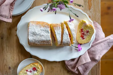 Eine köstliche Erdbeer Biskuitrolle, mit Puderzucker bestäubt und mit frischen Erdbeeren gefüllt, liegt auf einer weißen Platte.