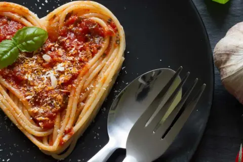 Herzförmige Pasta-Gerichte mit Tomatensoße, Basilikum und Parmesan. Daneben liegen ein Löffel, eine Gabel, Knoblauch und Tomaten.