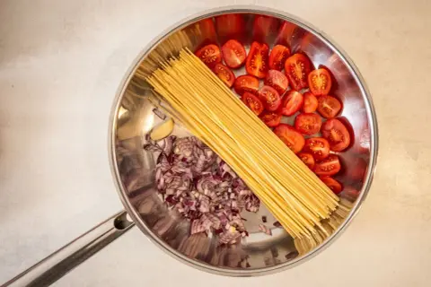 Zutaten für ein One-Pot-Gericht: Spaghetti, gehackte rote Zwiebeln, Kirschtomaten und eine Knoblauchzehe in einer Pfanne.