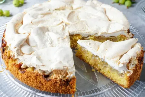 Ein saftiger Stachelbeer Baiser Kuchen mit goldbraunem Boden und einer luftigen Baiserhaube, ein Stück ist bereits angeschnitten.