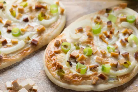 Zwei knusprige, vegetarische Flammkuchen mit Zwiebeln, Frühlingszwiebeln und würzigen Tofuwürfeln, bestreut mit etwas Mehl.