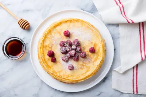 Ein Stapel goldbrauner Ofenpfannkuchen, garniert mit Himbeeren und Puderzucker. Daneben eine Schale Honig und ein Honiglöffel. Perfekt für den Thermomix!