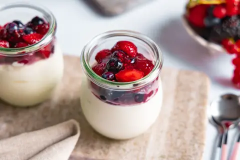 Zwei Gläser mit cremigem Dessert, gekrönt von Beeren und Fruchtsauce. Ein köstliches joghurtbombe rezept für den Sommer.