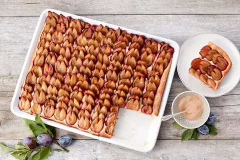 Ein köstlicher Zwetschgenkuchen vom Blech, mit saftigen Pflaumen belegt und goldbraun gebacken. Daneben ein Stück Kuchen auf einem Teller.