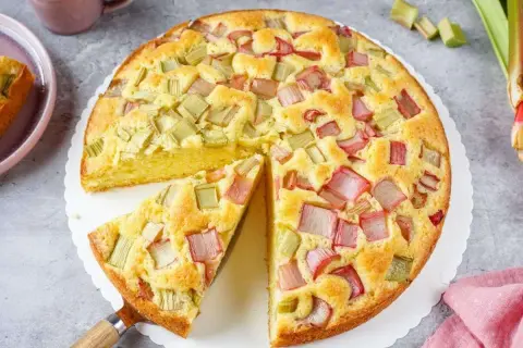 Saftiger Rhabarberkuchen in Springform, mit rosa und grünen Rhabarberstücken belegt, ein Stück herausgeschnitten.