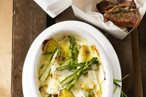 Überbackener Spargel mit Kartoffeln und Parmesan in einer Auflaufform, daneben ein Stück Lammkotelett.