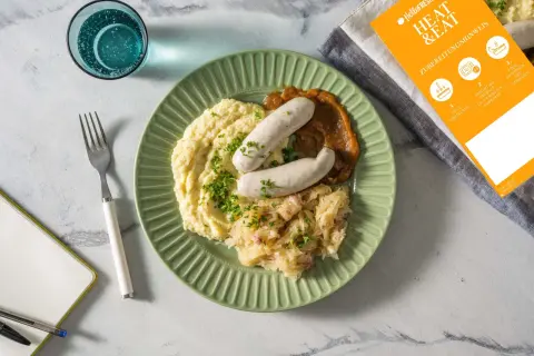 Fertig zubereitete Weißwurst mit Kartoffelpüree und Sauerkraut auf einem grünen Teller. Anleitung zum Weißwurst kochen liegt daneben.