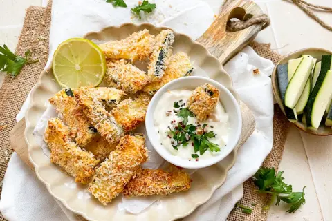 Knusprige Zucchini Pommes, paniert und goldbraun gebacken, serviert mit Dip und Limette.