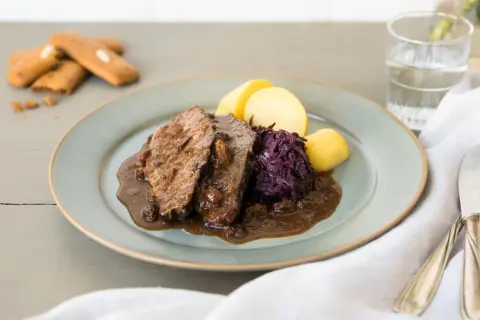 Ein Teller mit deftigem Braten, Rotkohl und Kartoffeln – ein schnelles Abendessen, das sättigt. Dazu ein Glas Wasser und ein paar Kekse.