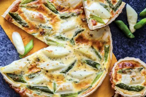 Herzhafte Quiche mit Spargel, goldbraun gebacken, auf einem orangen Tuch. Ein Stück ist angeschnitten, daneben liegen frische Spargelstücke.