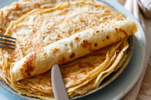 Ein Stapel goldbrauner Pfannkuchen, einer davon aufgerollt, liegt auf einem Teller. Ein Messer und eine Gabel liegen daneben. Perfekt für ein Pfannkuchen Rezept!